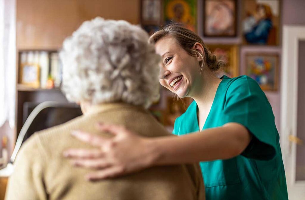 A senior caregiver helps a resident wash hands at a clean communal sink in a bright, modern assisted living facility.