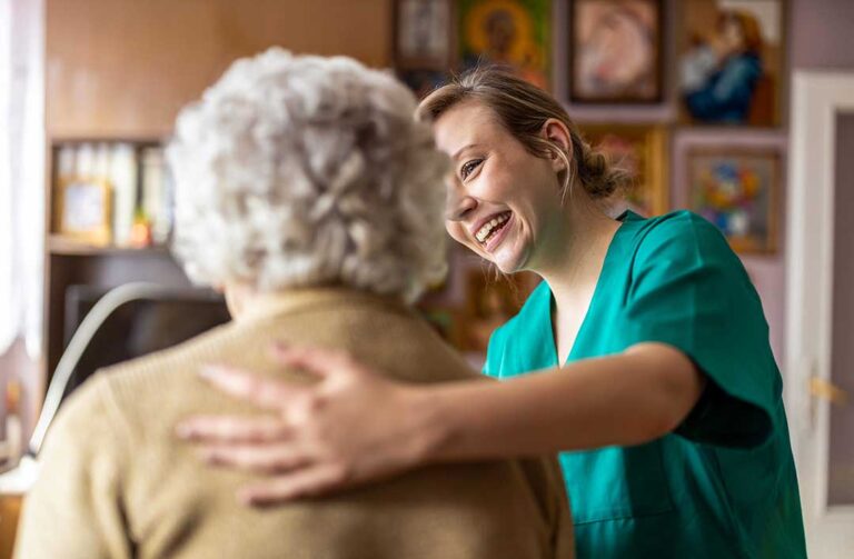 A senior caregiver helps a resident wash hands at a clean communal sink in a bright, modern assisted living facility.